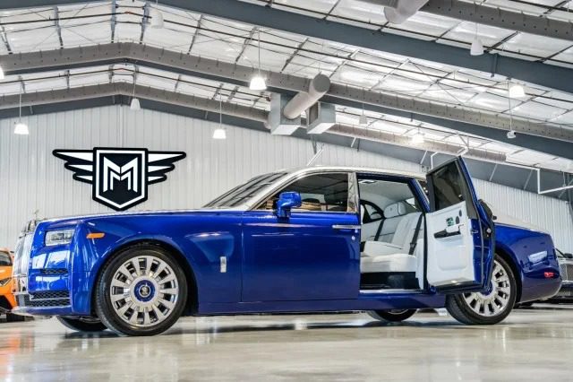 The quintessential luxury of a blue Rolls-Royce Phantom is showcased indoors, with one door open to reveal a pristine white interior. A black and white emblem graces the wall, completing this elegant scene.