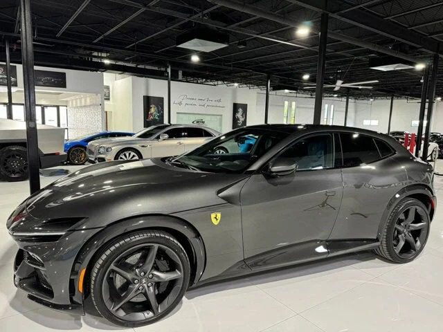 A sleek gray sports car parked indoors in a luxurious showroom, surrounded by super SUVs and other cars visible in the background.