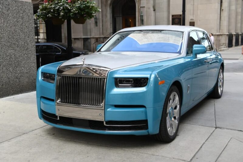 A quintessential luxury sedan, the blue Rolls-Royce Phantom is parked on a city street, showcasing its prominent chrome grille and sleek design.