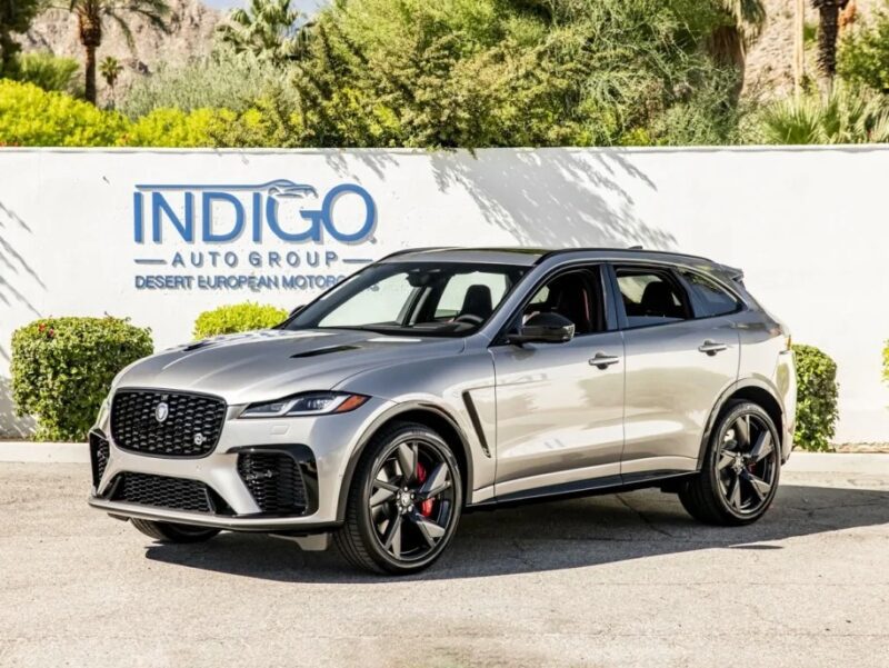 A luxury silver SUV is parked outdoors, showcasing its sleek design in front of a wall with "INDIGO AUTO GROUP DESERT EUROPEAN MOTORCARS" signage.