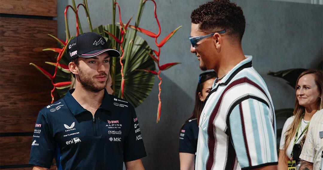 Two men stand and talk indoors at the Miami Grand Prix—one in a racing team shirt and cap, the other in sunglasses and a striped shirt. Three people are visible in the background during this F1 2024 Celebrity Sightings moment.