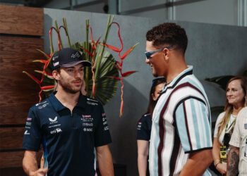 Two men stand and talk indoors at the Miami Grand Prix—one in a racing team shirt and cap, the other in sunglasses and a striped shirt. Three people are visible in the background during this F1 2024 Celebrity Sightings moment.