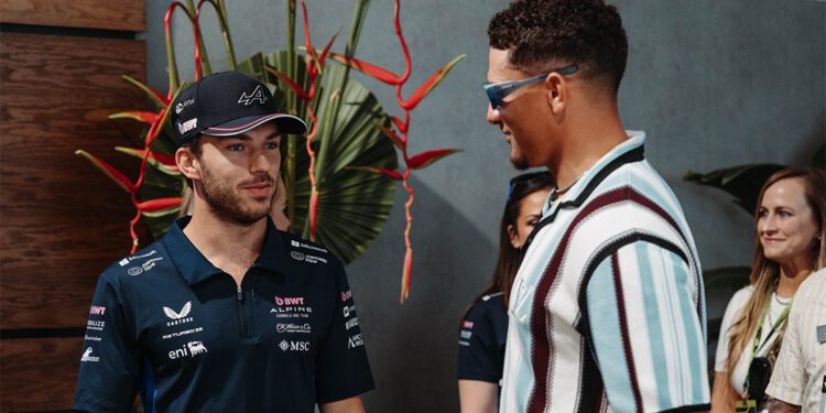 Two men stand and talk indoors at the Miami Grand Prix—one in a racing team shirt and cap, the other in sunglasses and a striped shirt. Three people are visible in the background during this F1 2024 Celebrity Sightings moment.