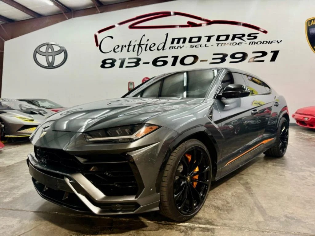 A gray Lamborghini Urus SUV, inspired by the fighting bull legacy, is parked indoors at a car dealership with Certified Motors signage and a large phone number on the wall behind it.