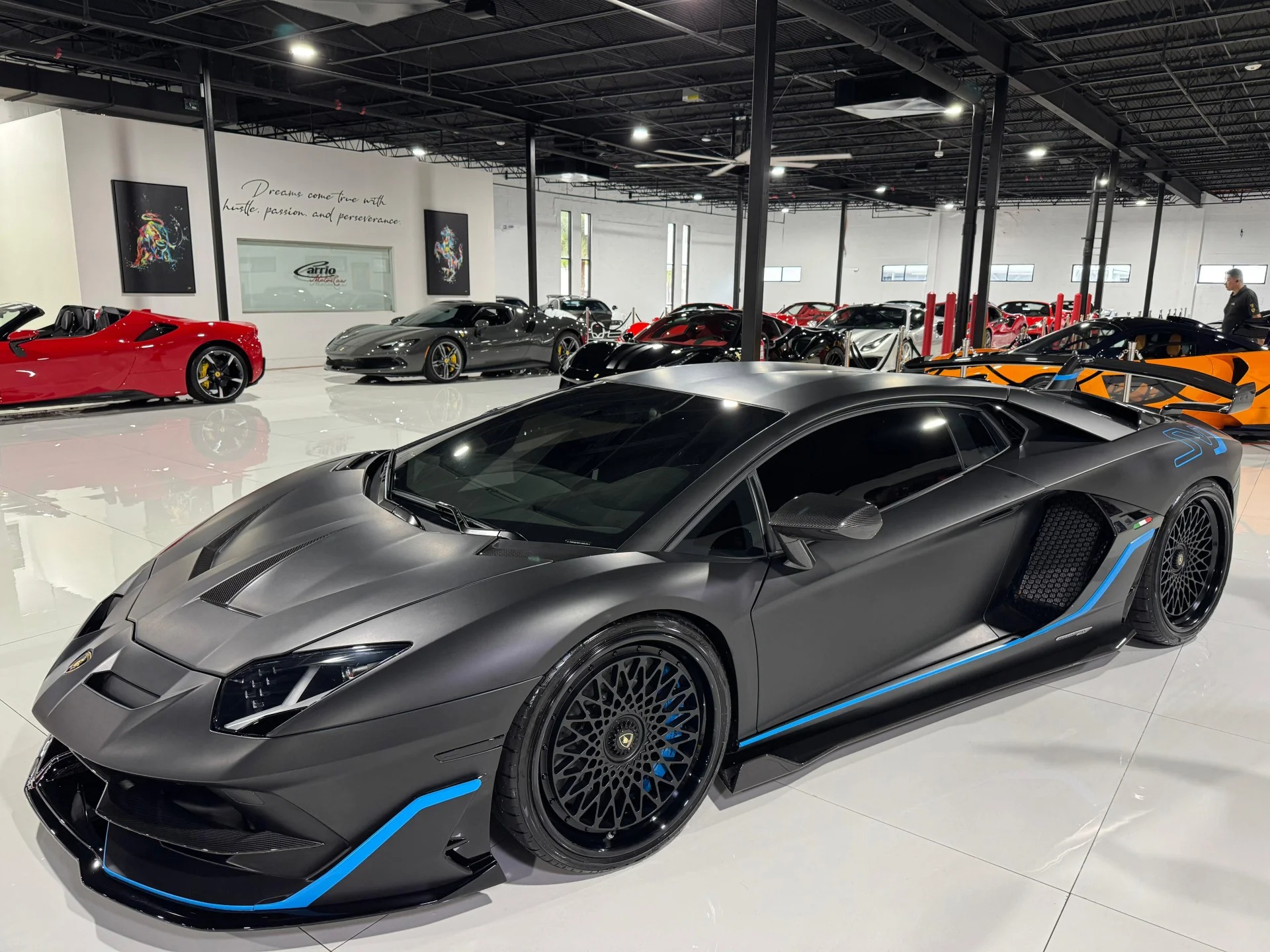 A matte black Lamborghini supercar with blue accents is parked in a showroom surrounded by various luxury sports cars under bright lights.