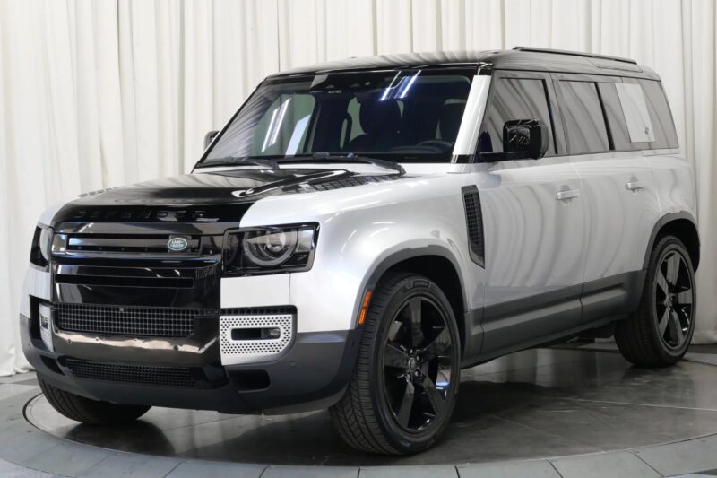Sidabrinis ir juodas „Land Rover Defender“ visureigis, vienas iš geriausiai prieinamų prabangių visureigių, yra pastatytas patalpose priešais baltos spalvos užuolaidų foną.