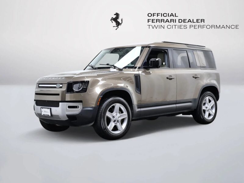 „Beige Land Rover Defender“, vienas iš geriausiai prieinamų prabangių visureigių, eksponuojamas studijos aplinkoje su "Oficialus „Ferrari“ pardavėjo „Twin Cities“ pasirodymas" Logotipas ant sienos už jos.