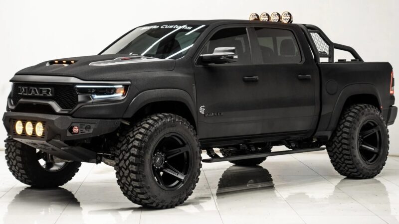 Matte black lifted luxury truck with large off-road tires, roof lights, front bumper lights, and custom grille showcases both capability and style, parked indoors on a glossy white floor.