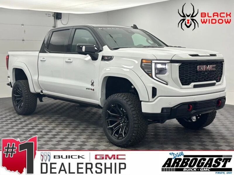 A white GMC Sierra AT4 Black Widow edition luxury truck is parked indoors on a checkered floor at a Buick GMC dealership, showcasing its impressive capability and bold presence.