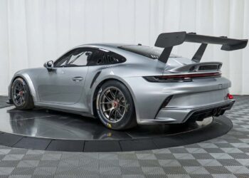 A silver Porsche race car with a large rear wing is displayed on a turntable in the showroom at Isringhausen Imports, highlighting its race-ready appeal against a checkered floor and white curtain backdrop—perfect for Porsche track cars enthusiasts.