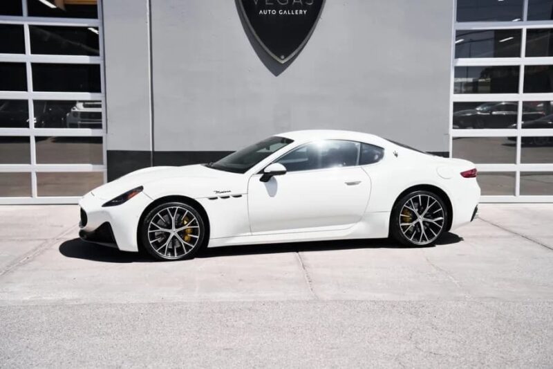 A white Maserati GranTurismo coupe, an icon of Italian luxury, is parked in front of a modern building with glass garage doors.