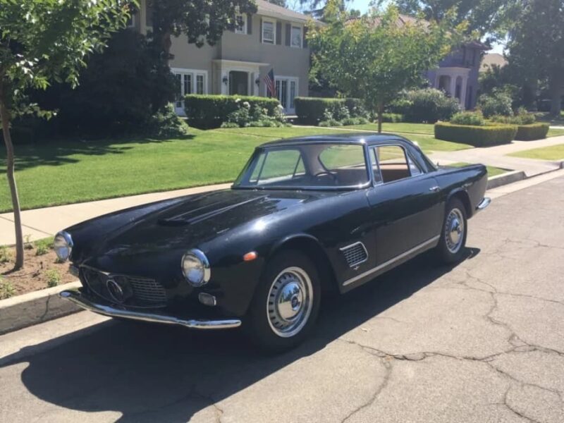 A classic black Maserati coupe parked on a residential street with houses, trees, and lawns in the background on a sunny day—Italian luxury at its finest.