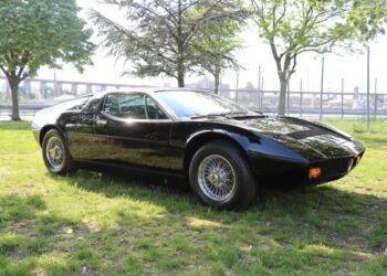 A black classic Maserati, a symbol of Italian luxury, is parked on grass under trees with a fence and bridge in the background.