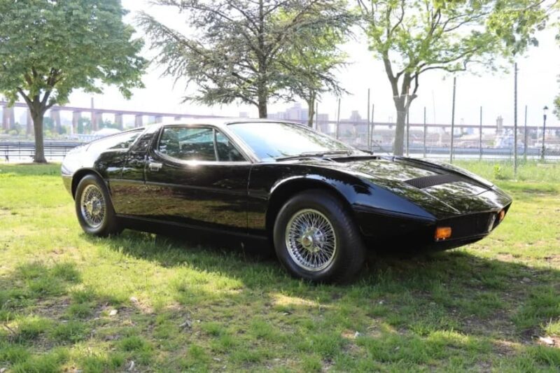 A black classic Maserati, a symbol of Italian luxury, is parked on grass under trees with a fence and bridge in the background.
