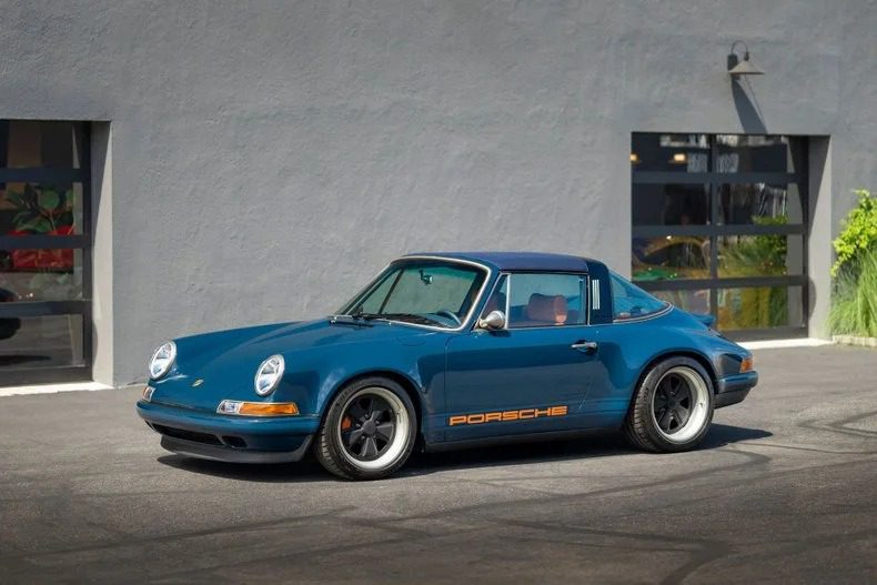 Senovinis mėlynas „Porsche 911“, primenantis legendinę oru aušinamų 911 erą, išdidžiai demonstruoja "Porsche" logotipas jo šone. Jis yra elegantiškai pastatytas priešais pilką pastatą su dideliais langais.