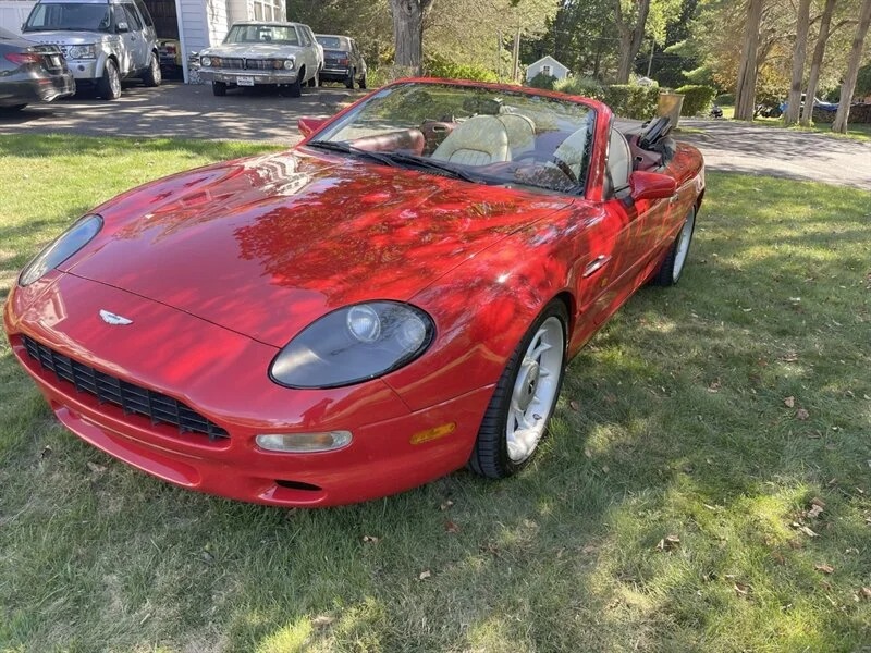 A red Aston Martin DB7 convertible, the epitome of British luxury, is parked on grass near a driveway, with other vehicles and trees in the background.