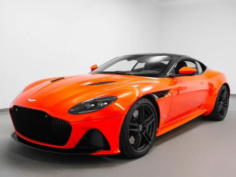 A bright orange Aston Martin sports car with a black roof and black wheels is parked indoors against a plain white background, showcasing true British luxury.