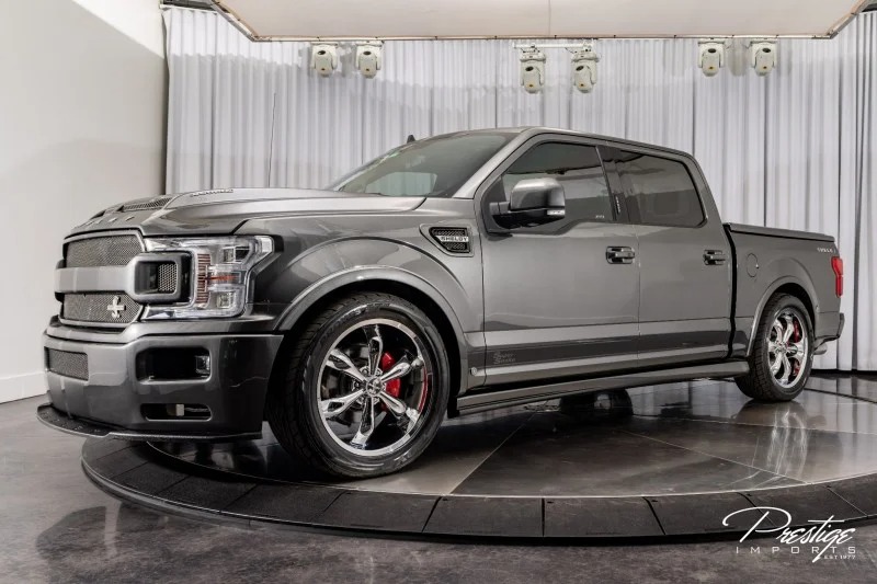 The sleek Ford F-150 features a sporty design with custom Shelby Trucks wheels, elegantly showcased indoors on a circular platform against a draped backdrop. Buy today and elevate your driving experience.