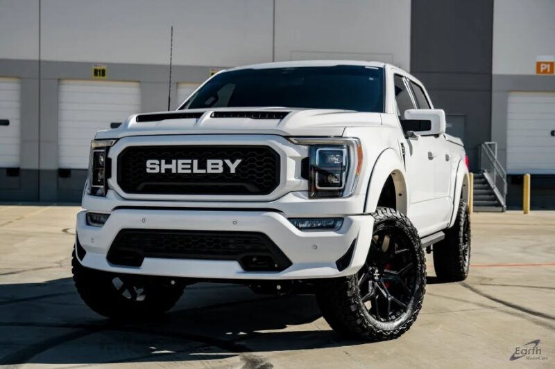 A white Shelby pickup truck, a stunning variant of the Ford F-150, is parked on a concrete surface in front of an industrial building with closed garage doors.