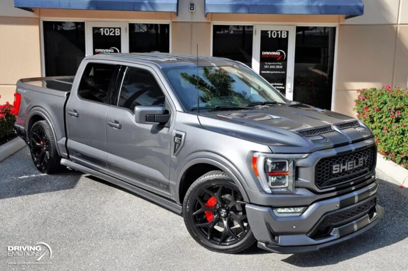 A sleek Ford F-150 with black and red details is parked outside a building, showcasing its connection to the legendary Shelby Trucks. Its bold presence next to the glass doors and plant beds invites you to buy today.