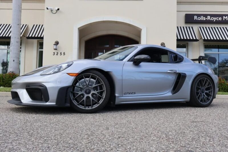 A sleek silver Porsche 718 sports car sits elegantly on the street, right in front of a Rolls-Royce dealership, with the model number GT4RS boldly visible on its side. This stunning vehicle is now available for sale to those who appreciate high-performance engineering.