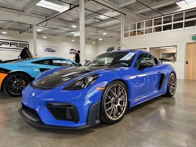 A Porsche 718 Cayman GT4 RS with a sleek black hood and striking bronze wheels sits in the garage, flanked by other vehicles.