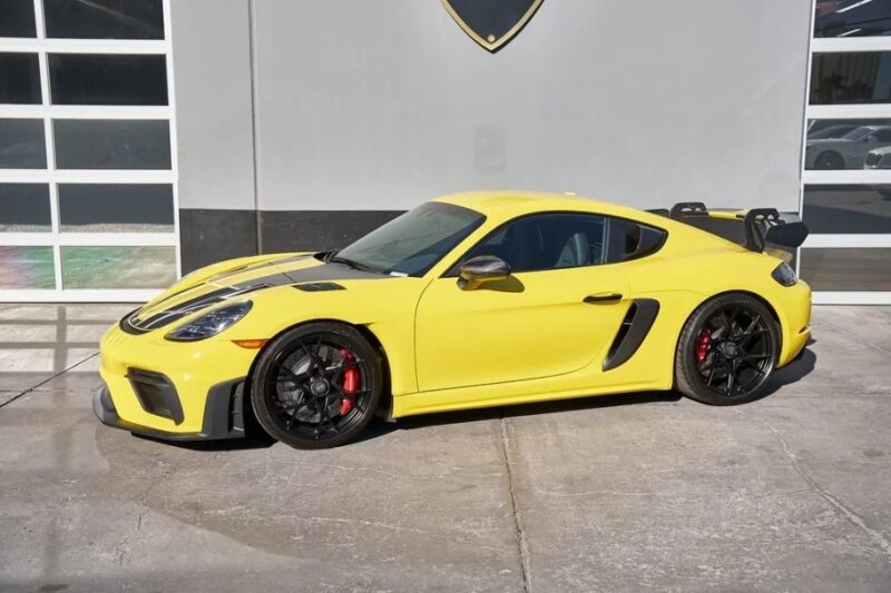 A sleek yellow Porsche 718 Cayman GT4 RS with black accents and a rear spoiler is parked in front of a garage with glass doors. This beauty is for sale, awaiting its next adventure on the open road.