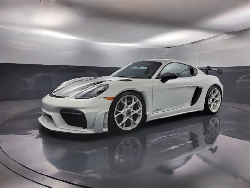 A white Porsche 718 Cayman GT4 RS with a sleek design and large rear spoiler is displayed in a well-lit showroom, available for sale.