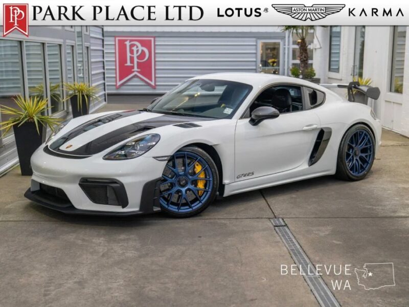 A white sports car, possibly a Porsche 718 Cayman GT4 RS, with striking blue wheels is parked for sale in front of Park Place Ltd in Bellevue, WA. Logos for Lotus, Aston Martin, and Karma are proudly displayed nearby.