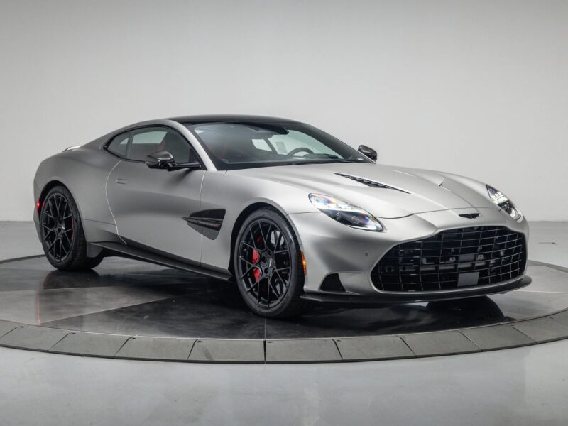 A silver Aston Martin coupe, a true performance machine with black wheels and red brake calipers, is displayed in a studio with a white background.