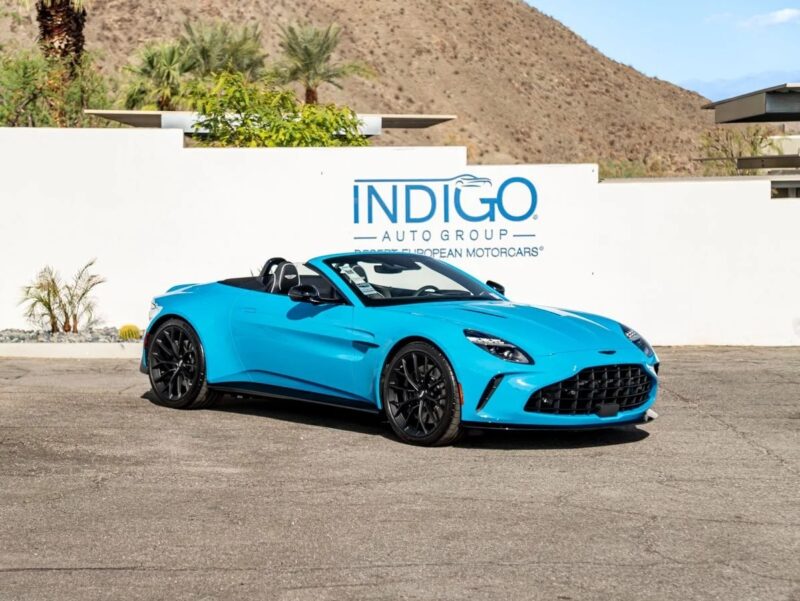 A blue Aston Martin convertible sports car is parked in front of a white wall with the "Indigo Auto Group" logo and palm trees in the background, showcasing true British luxury.