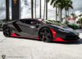 A sleek black and red 2017 Lamborghini sports car is parked outdoors, surrounded by palm trees and other vehicles, capturing the vibrant allure reminiscent of a Lamborghini Centenario.