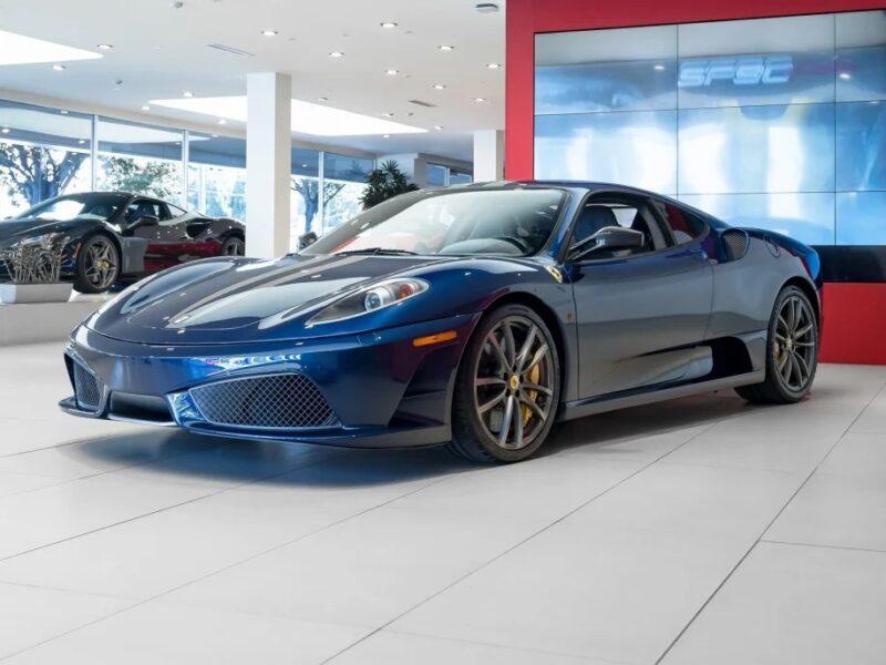 A blue Ferrari sports car is displayed in a showroom with large windows and a red accent wall. Other cars are visible in the background, showcasing an array of colors for sale.