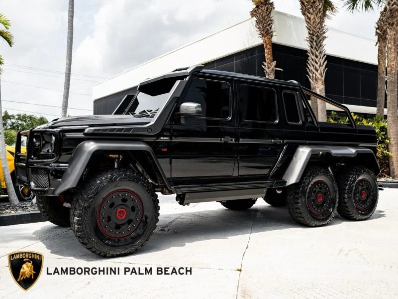 A black Mercedes-Benz G-Class 6x6 parked outside a modern building with palm trees is sure to turn heads. The Lamborghini Palm Beach logo is visible in the corner, enhancing the allure of this unique 6x6 truck.