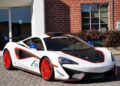 A white supercar with red accents and wheels is parked on a cobblestone driveway in front of a brick building and a glass door, hinting at its undeniable racing pedigree.