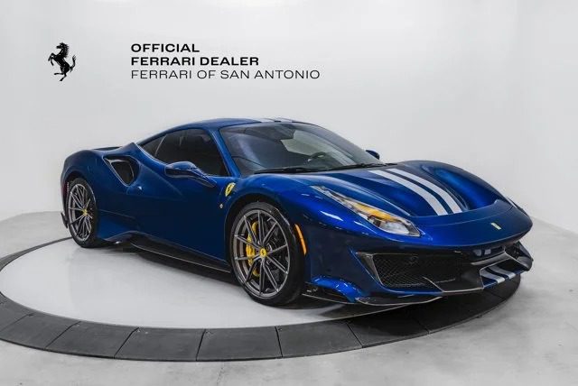 A blue Ferrari 488 Pista with white racing stripes is displayed in a showroom. The wall reads "Official Ferrari Dealer, Ferrari of San Antonio." This track-ready beast proudly showcases the iconic Prancing Horse emblem.