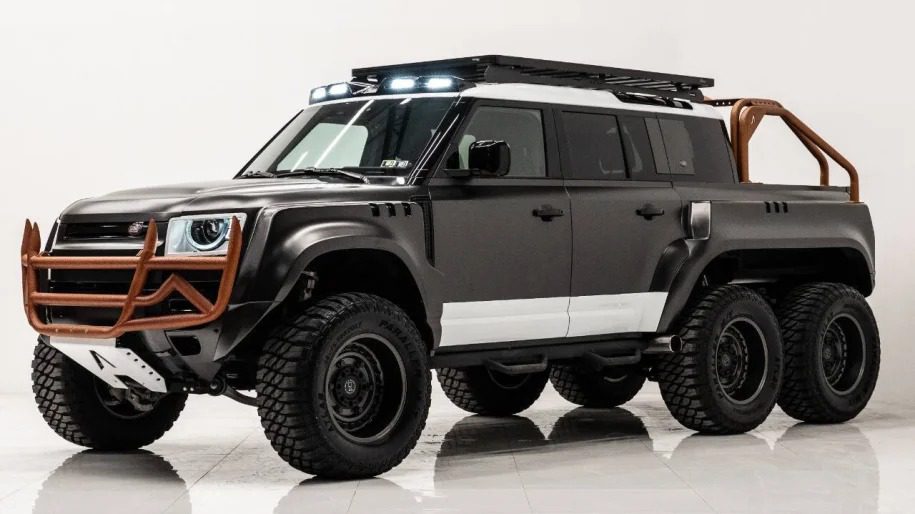 A custom black 6x6 SUV with a roof rack and rugged front bumper is parked indoors on a pristine white floor, designed to turn heads.