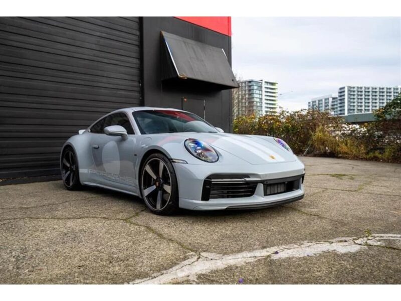 A silver Porsche 911 Sport Classic is parked on a paved surface near a black building, with modern apartment buildings visible in the background—a scene that beautifully reflects the enduring allure of Porsche history.