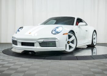 A white Porsche 911 Sport Classic with matching white wheels is displayed indoors on a turntable platform, set against a white curtain backdrop—a stunning nod to Porsche history.