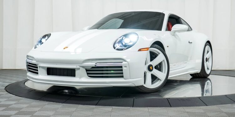 A white Porsche 911 Sport Classic with matching white wheels is displayed indoors on a turntable platform, set against a white curtain backdrop—a stunning nod to Porsche history.