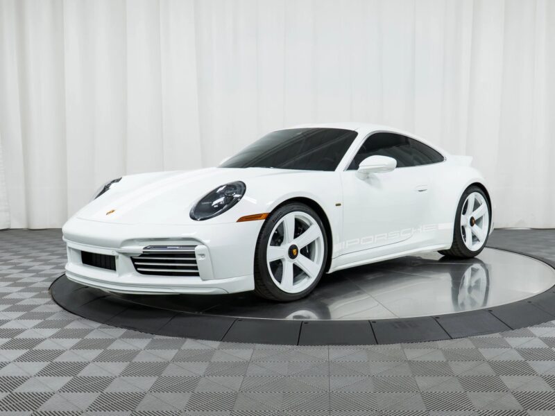 A white Porsche 911 coupe, a true icon in Porsche history, is displayed on a rotating platform with white wheels in a studio featuring a checkered floor and white curtains.