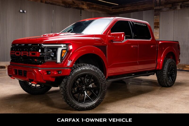 Displayed in a dimly lit showroom, the red Ford F-150 Raptor R boasts "FORD" on its grille and large black wheels. As a CARFAX 1-owner vehicle, it delivers impressive V8 performance, perfect for those seeking rugged off-road adventures.