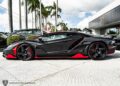 A sleek Lamborghini Centenario, adorned with striking red accents, basks in the sunshine. With palm trees swaying nearby and a dealership in view, this stunning sports car is proudly displayed for sale.