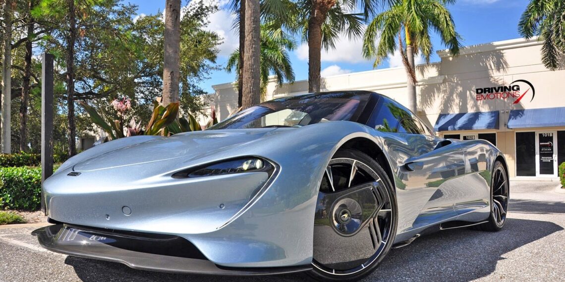 A sleek silver McLaren Speedtail sports car is parked in front of a building with palm trees, catching the eye of anyone interested in an exclusive sale.