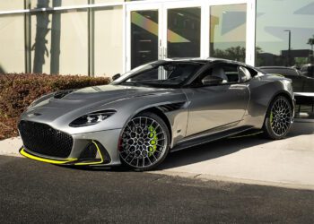 A silver sports car with neon yellow accents is parked in front of a modern glass building on a sunny day, featured in our Dealer Spotlight from Aston Martin Orlando, where exotics for sale redefine luxury and performance.
