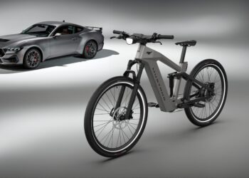 A sleek gray electric bike from the Ford eBikes collection stands in the foreground, while a gray sports car reminiscent of a Mustang looms in the background against a minimalist studio backdrop.