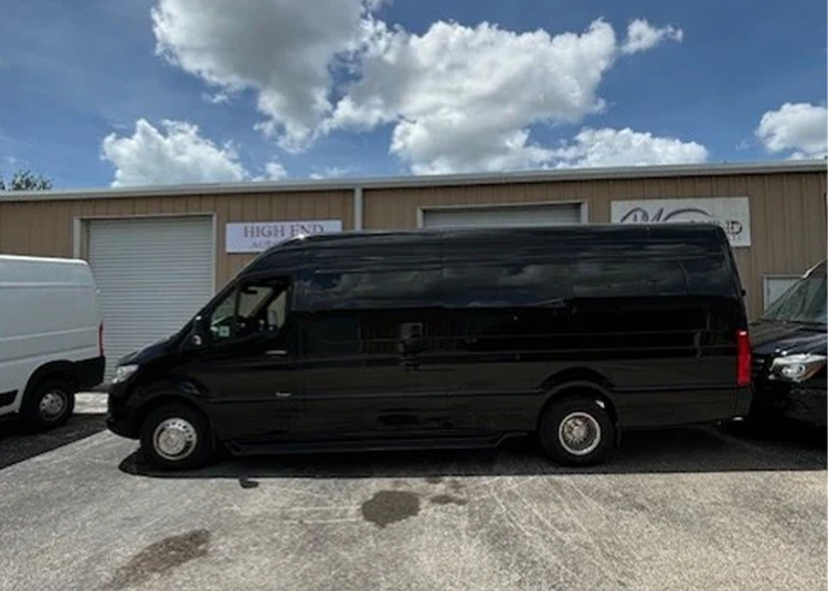 A large black Executive Sprinter van is parked outside a row of beige commercial garages on a sunny day with scattered clouds.