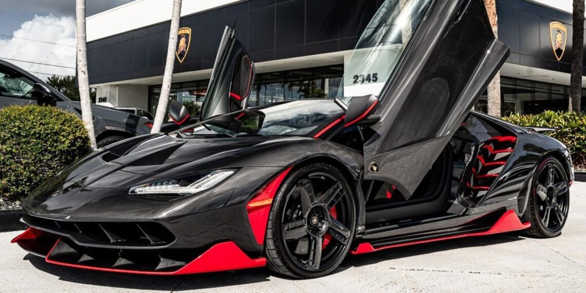 A 2017 Lamborghini Centenario in Giallo Midas with red accents and open scissor doors is parked in front of a dealership.