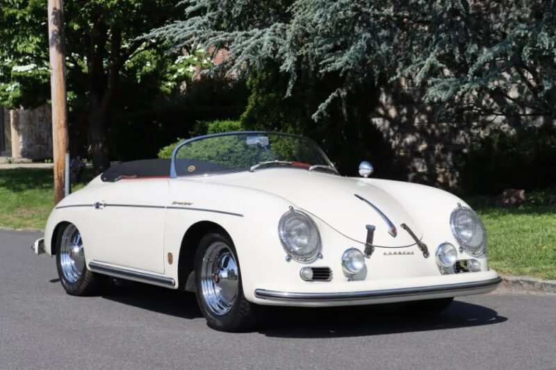 A vintage white Porsche 356 Speedster convertible, one of the best air-cooled Porsches, is parked on a suburban street in front of lush greenery and trees.