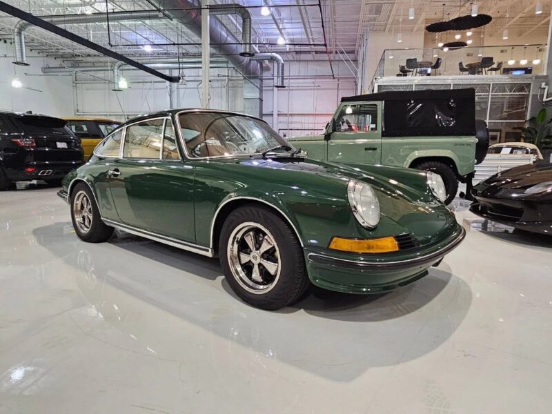 A classic green Porsche 911 coupe, one of the best air-cooled Porsches, is parked indoors in a well-lit showroom alongside other vehicles.